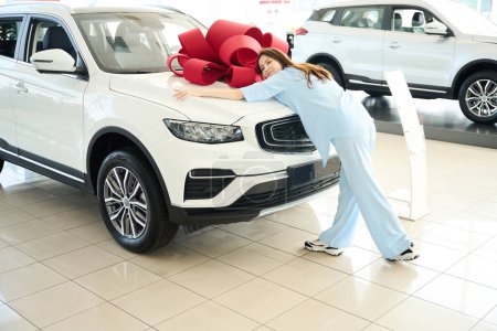 Foto de Encantadora dama abraza el capó de un coche con un arco de regalo, ella está en un concesionario de automóviles - Imagen libre de derechos