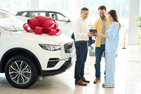 Buyers and a manager sign papers in a car dealership, a husband gives a car to his beloved wife