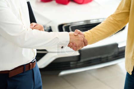Gerente felicita a un hombre en la compra de un coche, al lado de un coche con un arco de regalo rojo