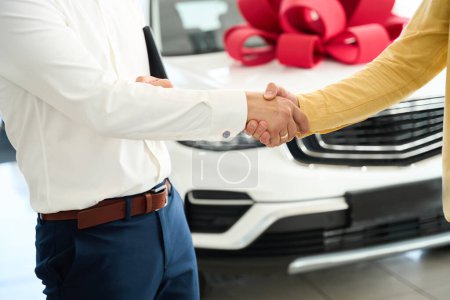 Téléchargez les photos : Le responsable du service client félicite un homme pour l'achat d'une voiture, à côté de la voiture avec un noeud cadeau rouge - en image libre de droit