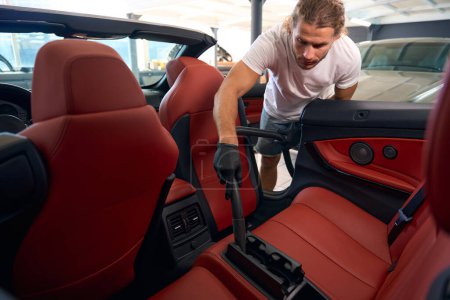 Photo for Young guy vacuums the interior of a car, he cleans the back seats - Royalty Free Image