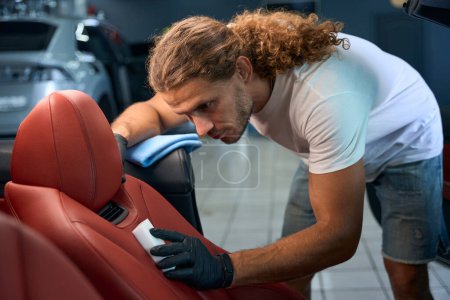 Téléchargez les photos : Jeune homme nettoie siège conducteur en cuir avec une éponge de mélamine, il fait des détails pour l'intérieur d'une voiture élégante - en image libre de droit