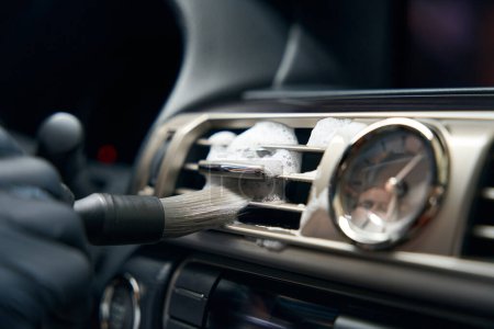 Foto de El hombre con guantes de protección limpia un conducto de aire del coche con un cepillo especial, hay un reloj en el salpicadero - Imagen libre de derechos