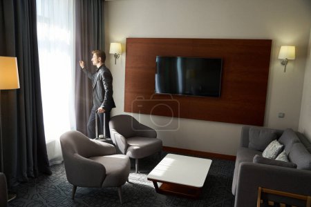 Téléchargez les photos : Homme en tenue de voyage se tient près de la fenêtre dans une chambre d'hôtel, la chambre a un design minimaliste moderne - en image libre de droit