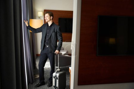 Foto de Sonriente en ropa de viaje se encuentra junto a la ventana en una habitación de hotel, la habitación tiene un diseño minimalista moderno - Imagen libre de derechos