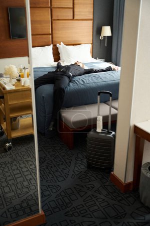 Foto de Hombre yace con los brazos extendidos en una cama grande en una habitación de hotel, él está en ropa de viaje negro - Imagen libre de derechos
