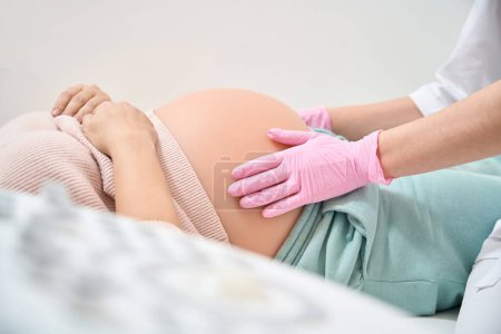 Foto de Ginecólogo-obstetra calificado en guantes médicos que examinan el vientre de la mujer en el segundo trimestre del embarazo, comprobando la presentación del bebé - Imagen libre de derechos