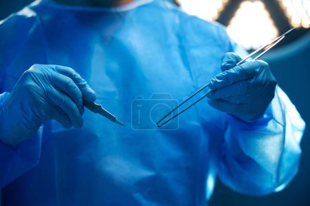 Téléchargez les photos : Chirurgien professionnel méconnaissable tenant une pince à épiler et un scalpel dans la salle d'opération - en image libre de droit