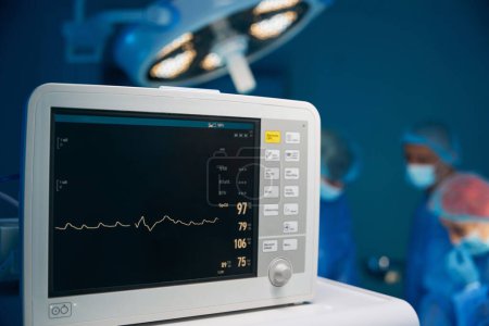 Foto de Electrocardiograma en la sala de urgencias de cirugía hospitalaria que muestra la frecuencia cardíaca del paciente con el equipo de cirujanos en el fondo - Imagen libre de derechos