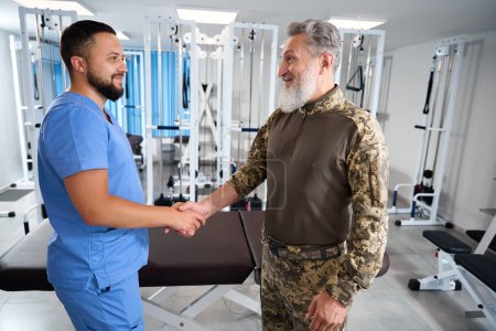 Foto de Quiropráctico saluda a un hombre con ropa militar en un centro de rehabilitación, los hombres se dan la mano - Imagen libre de derechos