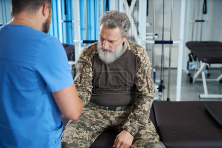 Foto de Médico de rehabilitación examina las heridas de un militar en un centro de rehabilitación, los hombres se encuentran en el gimnasio - Imagen libre de derechos