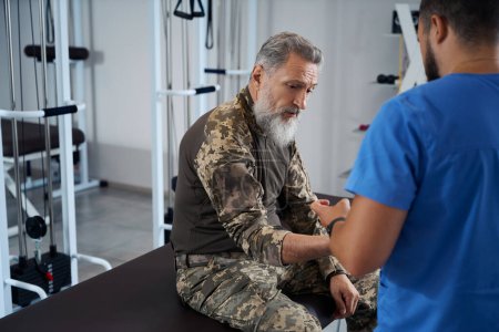 Doctor examina el brazo herido de un militar en un centro de rehabilitación, los hombres se encuentran en el gimnasio