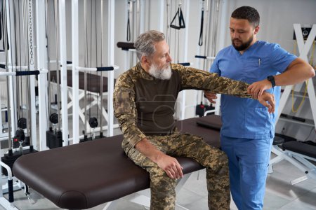 Foto de Barbudo vestido de militar se sienta en una mesa de masaje mientras es examinado por un fisioterapeuta - Imagen libre de derechos