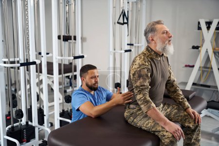 Photo for Chiropractor working on the back of a man in military clothing, patient sitting on massage table - Royalty Free Image