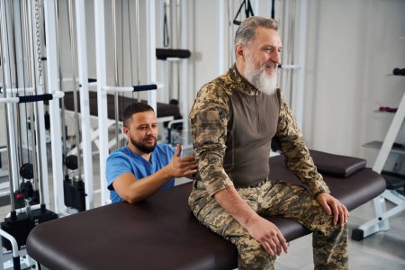 Photo for Physiotherapist working on the back of a man in military clothing, patient sitting on massage table - Royalty Free Image