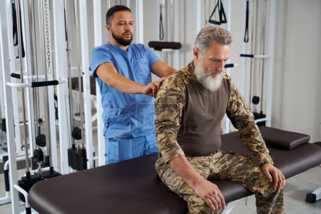 Foto de El médico examina la espalda de un hombre vestido de militar, el paciente se sienta en una mesa de masaje - Imagen libre de derechos