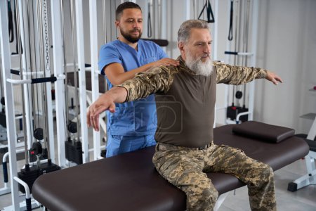 Photo for Rehabilitation therapist works with a patient in military clothing, a man sits on a massage table - Royalty Free Image
