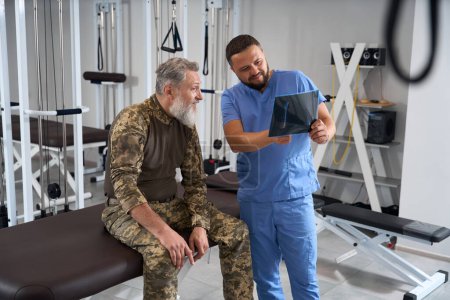 Foto de Fisioterapeuta con un paciente en ropa militar examina rayos X, ambos hombres son barbudos - Imagen libre de derechos