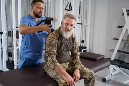 Foto de El médico lleva a cabo una sesión de masaje de espalda de hardware para un militar en rehabilitación, un paciente con ropa de camuflaje - Imagen libre de derechos