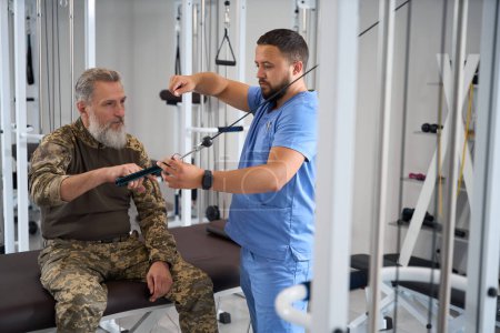 Foto de Terapeuta de rehabilitación ayuda a un paciente militar a trabajar en una máquina, un hombre hace ejercicios de estiramiento muscular - Imagen libre de derechos