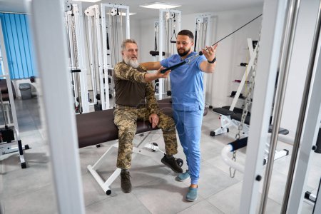 Foto de Entrenador ayudando al paciente militar a trabajar en la máquina de ejercicios, hombre haciendo ejercicios de estiramiento muscular - Imagen libre de derechos
