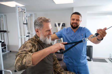 Foto de Especialista ayuda a un paciente militar a trabajar en una máquina, un hombre hace ejercicios de estiramiento muscular - Imagen libre de derechos