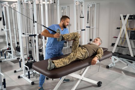 Military patient doing muscle stretching exercises in a rehabilitation center, he is being helped by an experienced doctor
