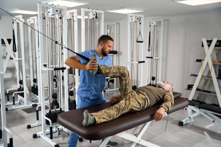 Foto de El paciente hace ejercicios de estiramiento muscular en un simulador especial, es ayudado por un rehabilitador experimentado - Imagen libre de derechos
