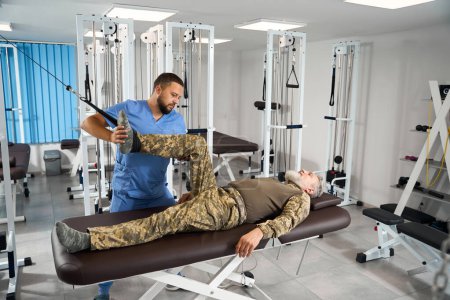 Photo for Man does exercises to stretch his leg muscles on a special simulator, he is helped by an experienced rehabilitator - Royalty Free Image