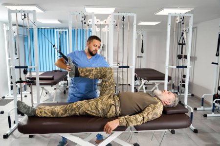 Photo for Bearded man does exercises to stretch his leg muscles on a special simulator, he is helped by an experienced rehabilitator - Royalty Free Image