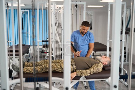 Foto de El paciente militar hace ejercicios para estirar los músculos de su brazo en un simulador especial, es ayudado por un rehabilitador - Imagen libre de derechos
