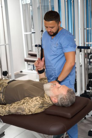 Téléchargez les photos : Physiothérapeute aide un patient à effectuer des exercices pour étirer les muscles des bras sur une machine spéciale - en image libre de droit
