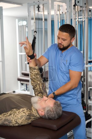Photo for Physiotherapist helps a military man perform exercises to stretch his arm muscles on a special machine - Royalty Free Image