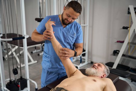 Foto de Especialista en rehabilitación examina una mano de hombre, el paciente se acuesta en una mesa de masaje - Imagen libre de derechos