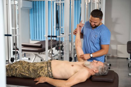 Foto de Especialista fisioterapeuta consultando a un hombre en ropa militar, el paciente se acuesta en una mesa de masaje - Imagen libre de derechos