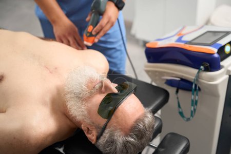 Doctor works with patients scars at laser therapy session, male in safety glasses lies on couch