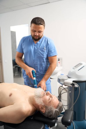 Téléchargez les photos : Thérapeute de réadaptation travaille dans une clinique avec un patient aux cheveux gris, un homme s'allonge sur un canapé avec un torse nu - en image libre de droit