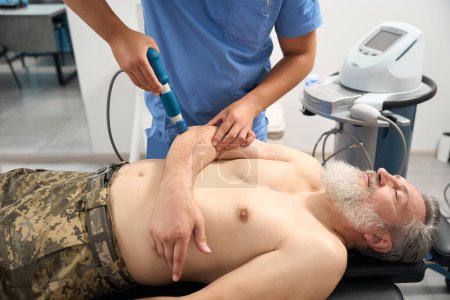 Téléchargez les photos : Le docteur fait une thérapie par ondes de choc à un patient militaire, un homme s'allonge sur un canapé avec un torse nu - en image libre de droit