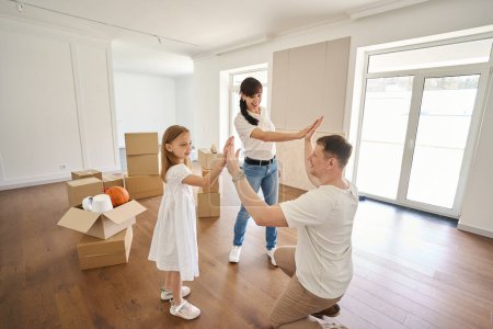 Photo for Father giving five to wife and daughter in new modern sunny townhouse during moving. Happy european parents and child. Family future planning, relationship and spending time together - Royalty Free Image