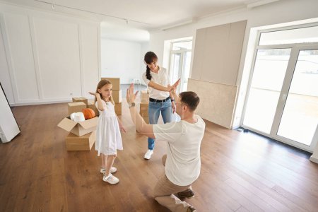 Photo for Father giving five to wife and daughter in new modern sunny townhouse during moving. Joyful caucasian parents and child. Family future planning, relationship and enjoying time together - Royalty Free Image