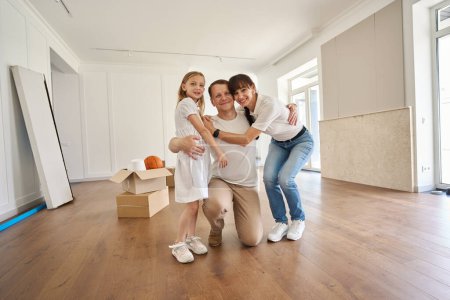 Photo for Happy caucasian mother, father and daughter hugging and looking at camera in new sunny modern townhouse during moving. Family future planning, relationship and enjoying time together - Royalty Free Image