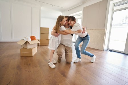 Photo for Pleased caucasian mother, father and daughter embracing and looking at each other from in sunny modern townhouse during moving. Family future planning, relationship and enjoying time together - Royalty Free Image