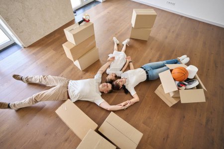 Photo for Top view of caucasian mother, father and daughter holding hands, looking up and dreaming on floor in new townhouse during moving. Family future planning, relationship and enjoying time together - Royalty Free Image