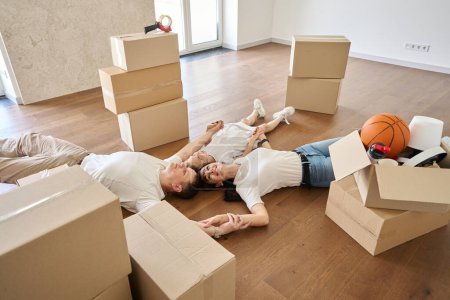 Photo for Beautiful caucasian mother, father and daughter holding hands and dreaming on floor in new sunny modern townhouse during moving. Family future planning, relationship and enjoying time together - Royalty Free Image