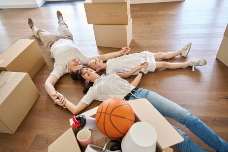 Photo for Caucasian mother, father and daughter holding hands, looking up and dreaming on floor in new sunny modern townhouse during moving. Family future planning, relationship and spending time together - Royalty Free Image