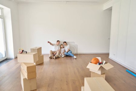 Photo for Man showing something to his wife and daughter while they sitting on floor in new modern townhouse during moving. Family future planning, relationship and spending time together - Royalty Free Image