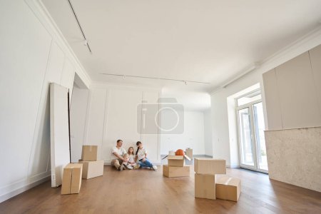 Photo for Focused european mother, father and daughter sitting on floor and looking at new modern townhouse during moving. Family future planning, relationship and spending time together - Royalty Free Image