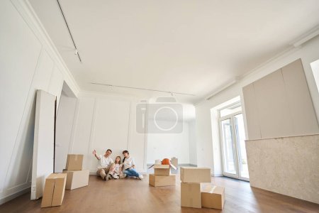 Photo for Father showing something to his wife and daughter while they sitting on floor in new modern townhouse during moving. Family future planning, relationship and spending time together - Royalty Free Image