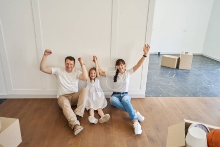 Photo for Pleased caucasian mother, father and daughter with hands in air sitting on floor, looking at camera and celebrating moving to new modern townhouse. Family future planning and spending time together - Royalty Free Image