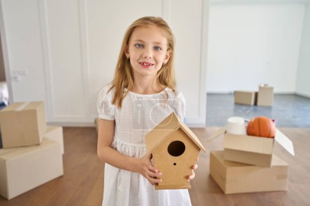 Foto de Sonriente niña caucásica sosteniendo pajarera de madera y mirando a la cámara en la nueva casa moderna borrosa durante el movimiento. Planificación futura de la vida - Imagen libre de derechos